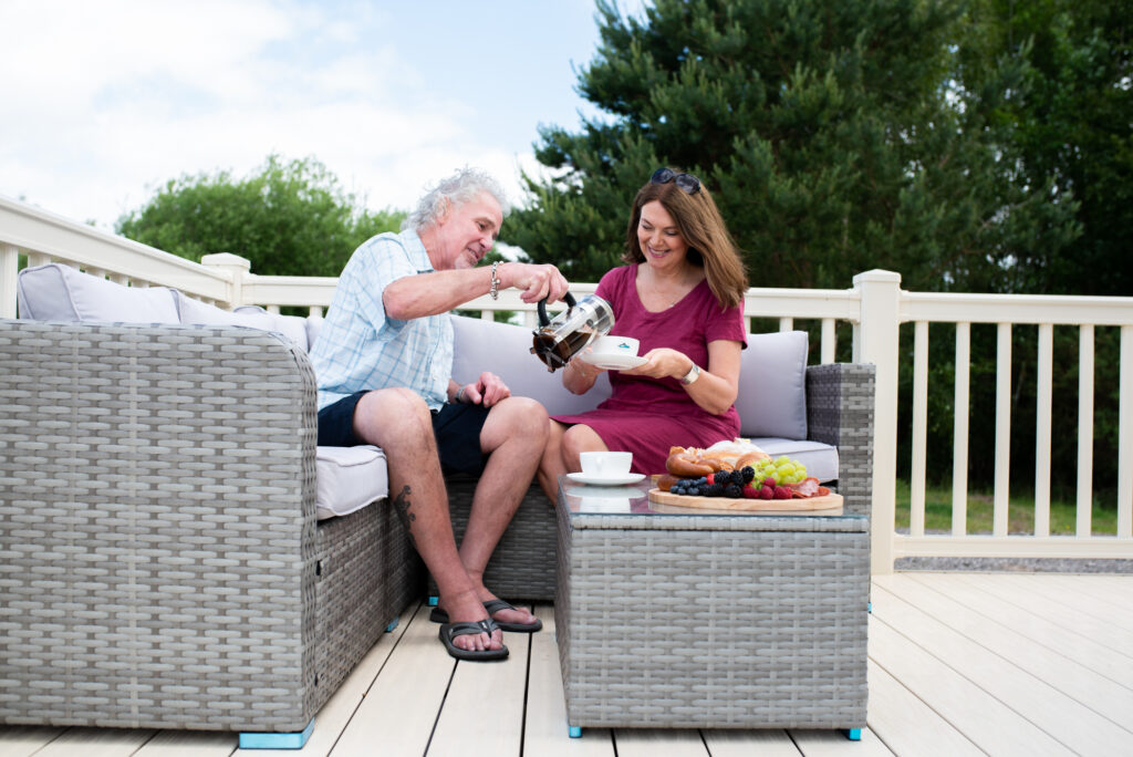 Couple sat on outdoor furniture on caravan decking, drinking coffee